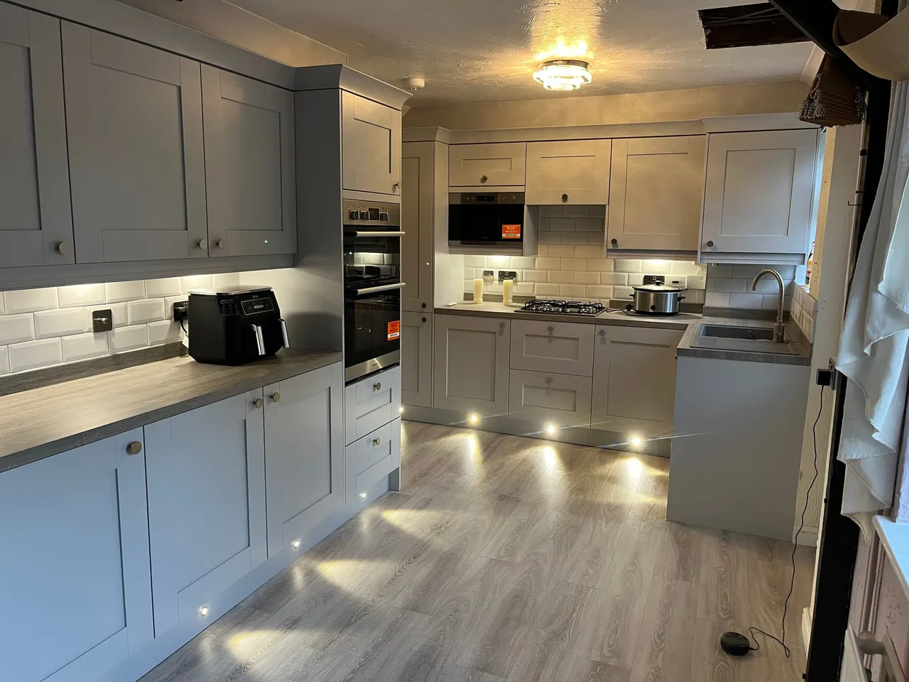 Grey shaker kitchen with wood-effect worktops, integrated oven, gas hob and plinth lighting over grey vinyl flooring