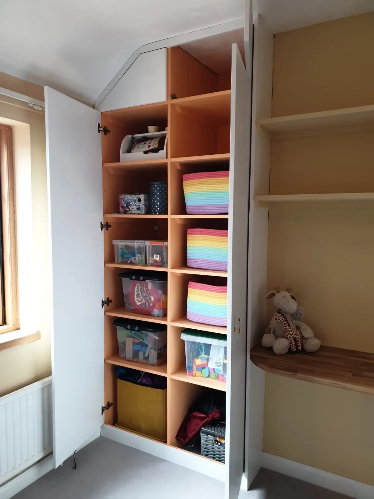 Fitted alcove cupboard with open cubby shelving holding storage baskets and toys, beside a built-in bench
