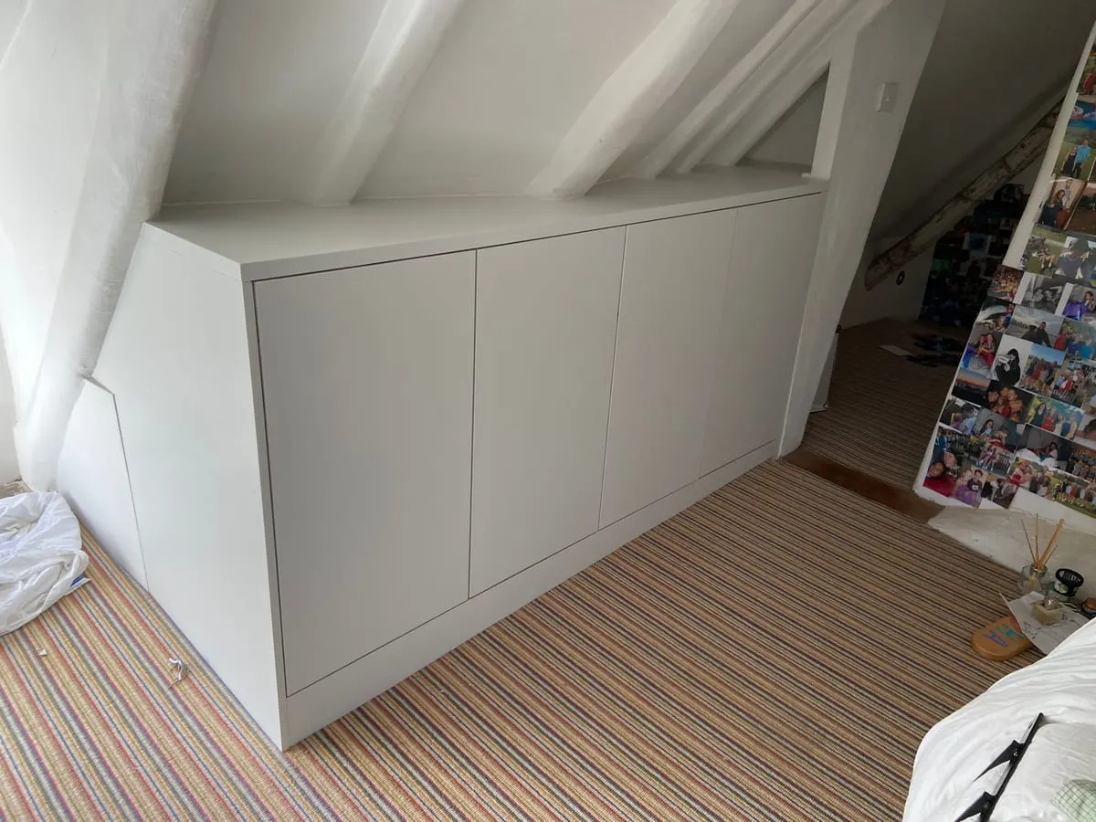 Built-in white handleless eaves cupboards fitted along a sloping loft bedroom wall with striped carpet