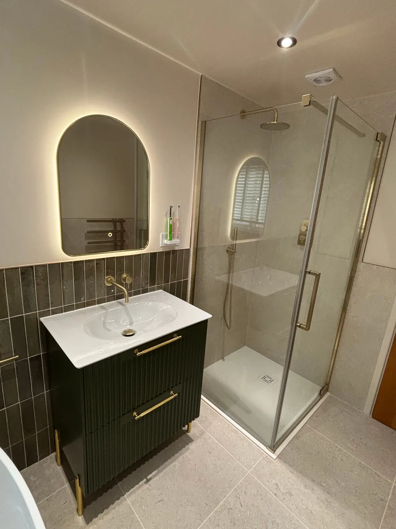 Green fluted vanity unit with brass taps, arched backlit mirror and glass walk-in shower enclosure