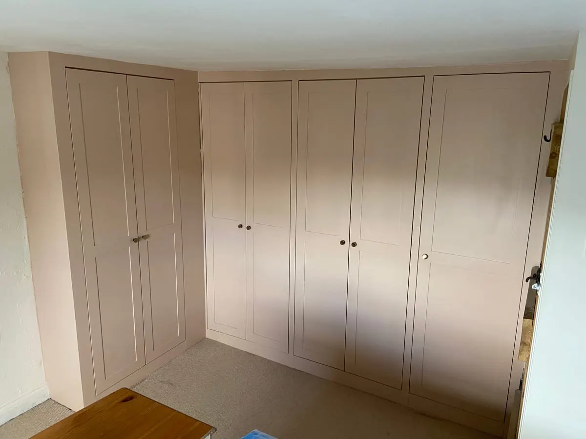 Floor-to-ceiling fitted wardrobes with pale pink shaker doors wrapping around a bedroom corner