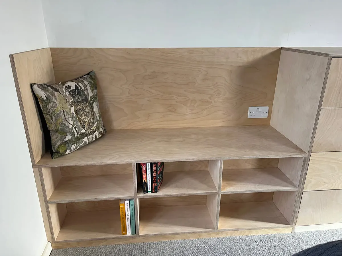 Birch plywood window seat with open cubby shelving beneath, holding books and a patterned cushion