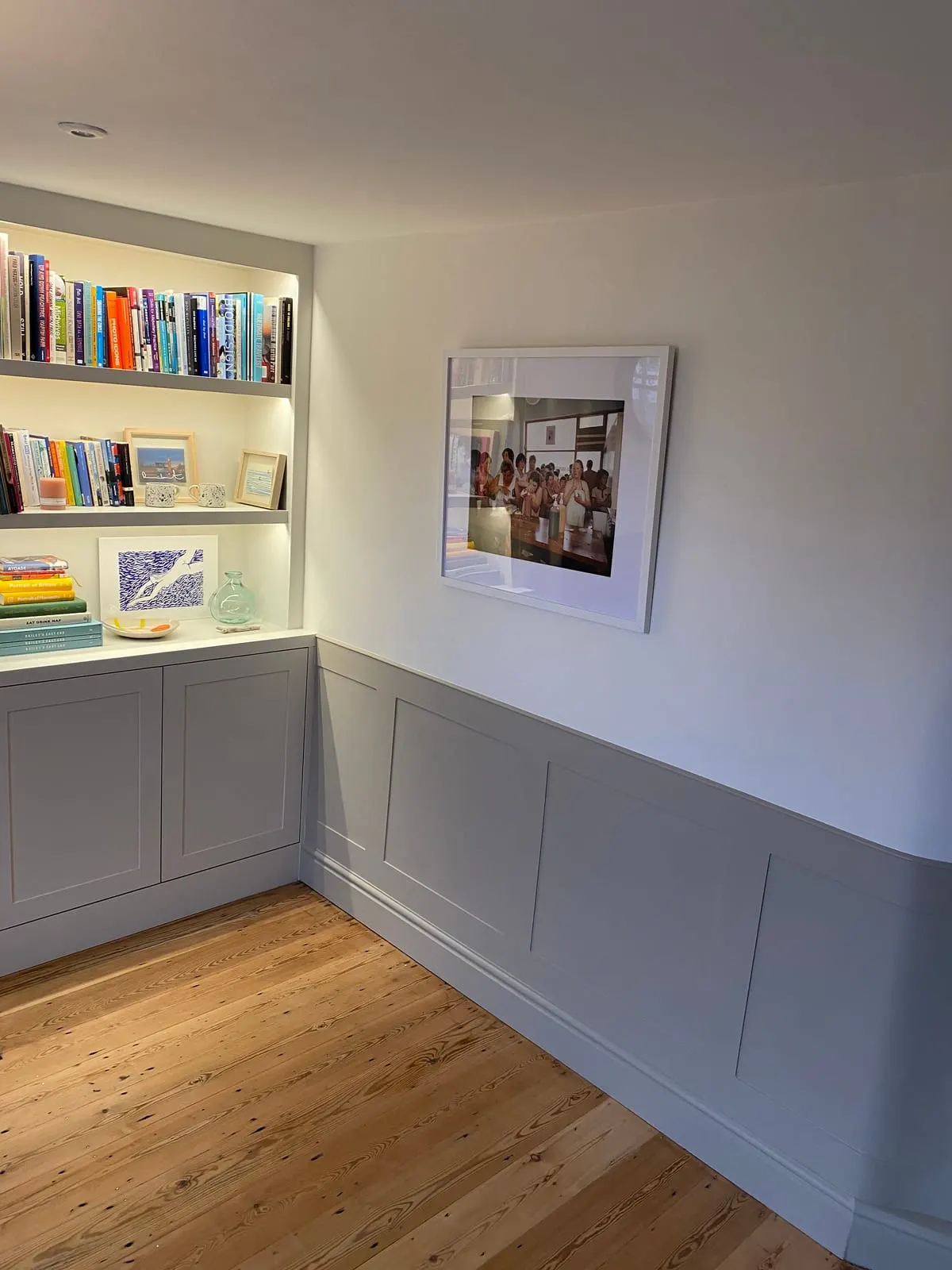 Grey fitted alcove shelving with lit book display above shaker cupboards, beside painted wall panelling