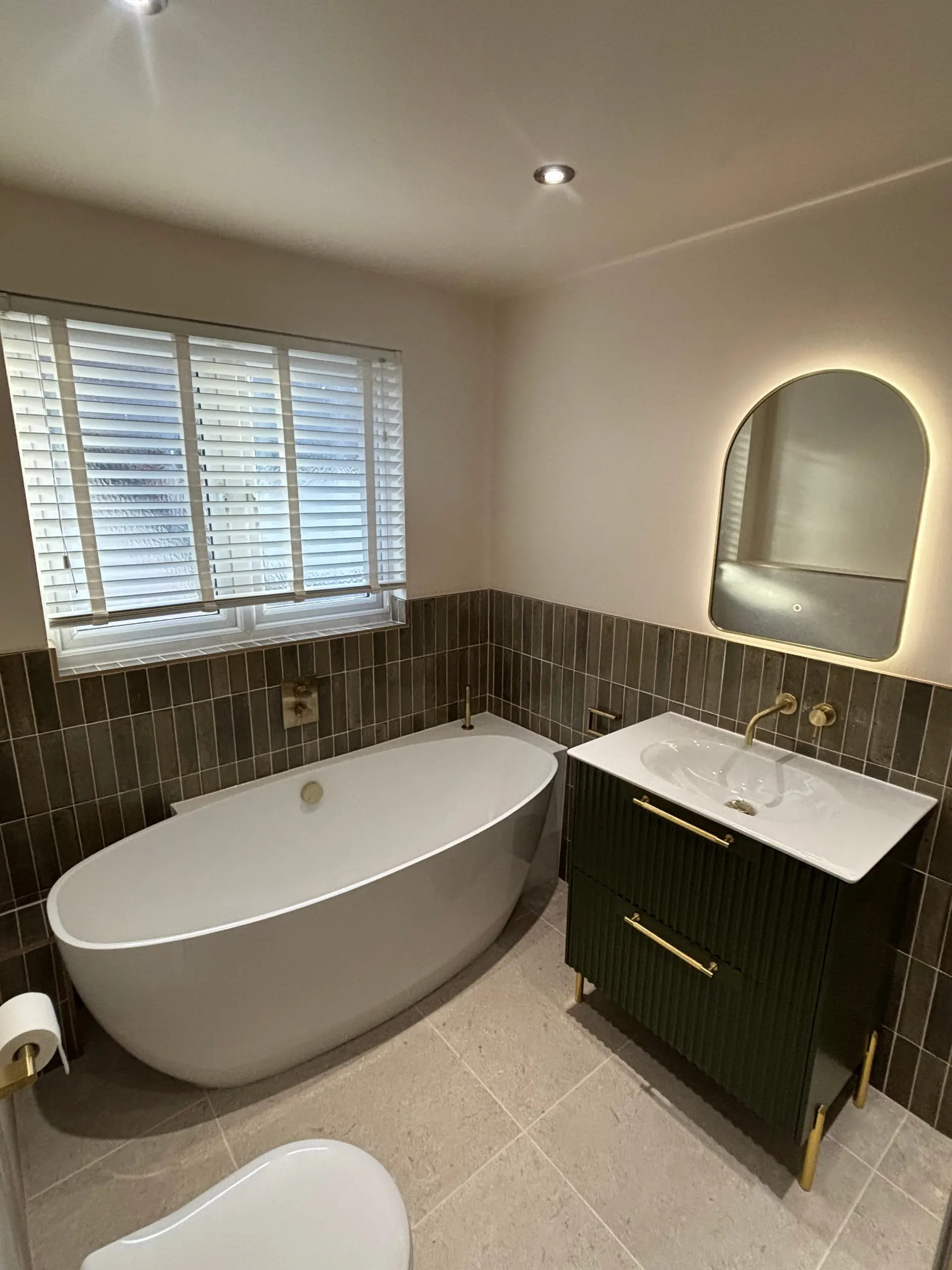 Freestanding white bath with green wall tiles, fluted green vanity unit, brass taps and arched backlit mirror