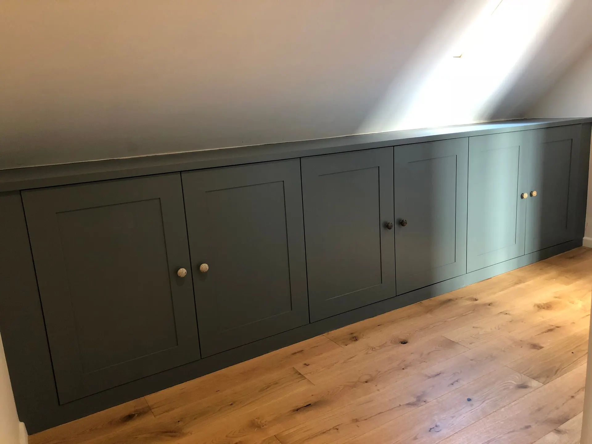 Long run of dark green shaker eaves cupboards with round knobs above a solid oak floor
