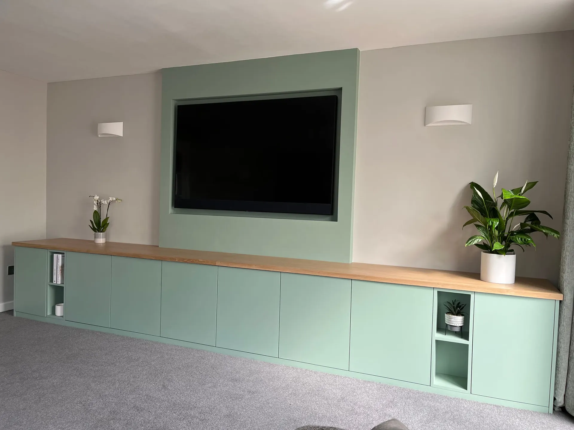 Full-width fitted sage green media unit with solid oak top and recessed TV panel above