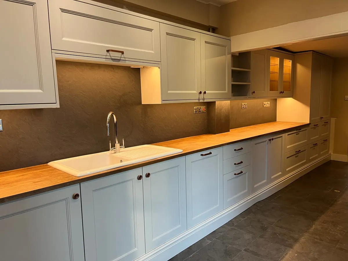 Long run of pale blue shaker kitchen cabinetry with oak worktops, ceramic sink and slate splashback