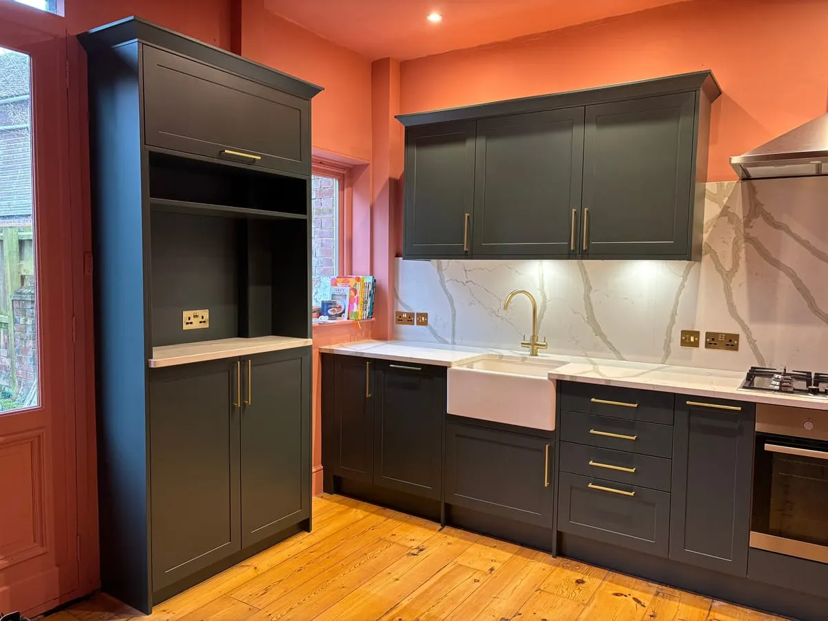 Dark green shaker kitchen with tall larder unit, marble-effect splashback, brass handles and Belfast sink