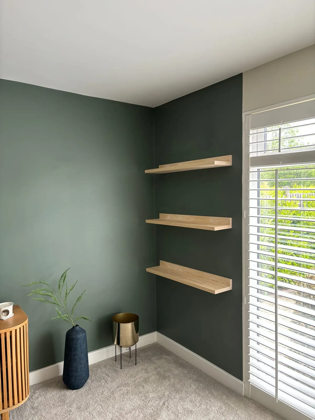 Three oak floating shelves fitted to a dark green wall beside a window with white shutters