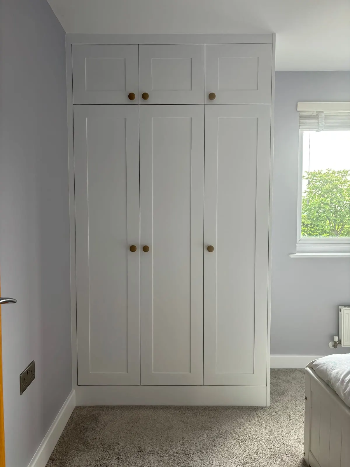 Floor-to-ceiling fitted white shaker wardrobes with brass knobs and overhead storage in a bedroom