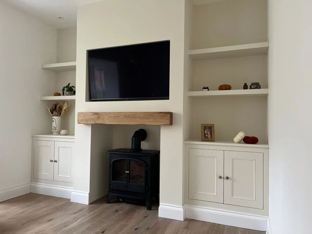 Cream fitted alcove cupboards and floating shelves either side of a log burner with oak beam mantel